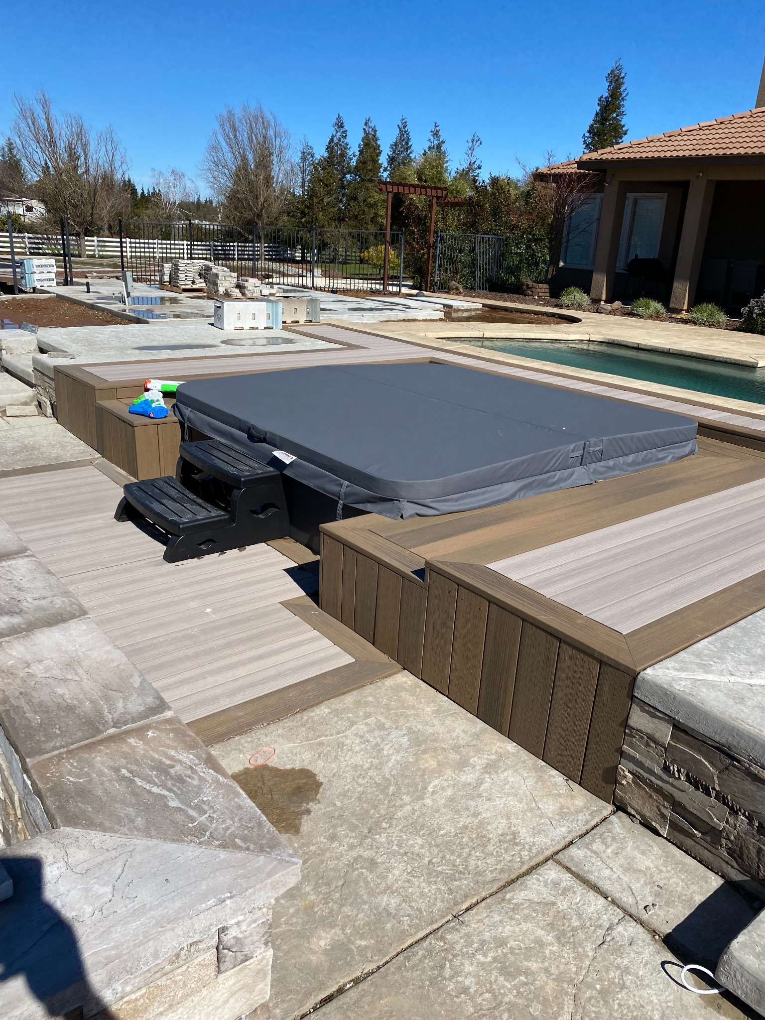 Decking surrounding a covered spa beside a swimming pool, concrete paving and landscaping.