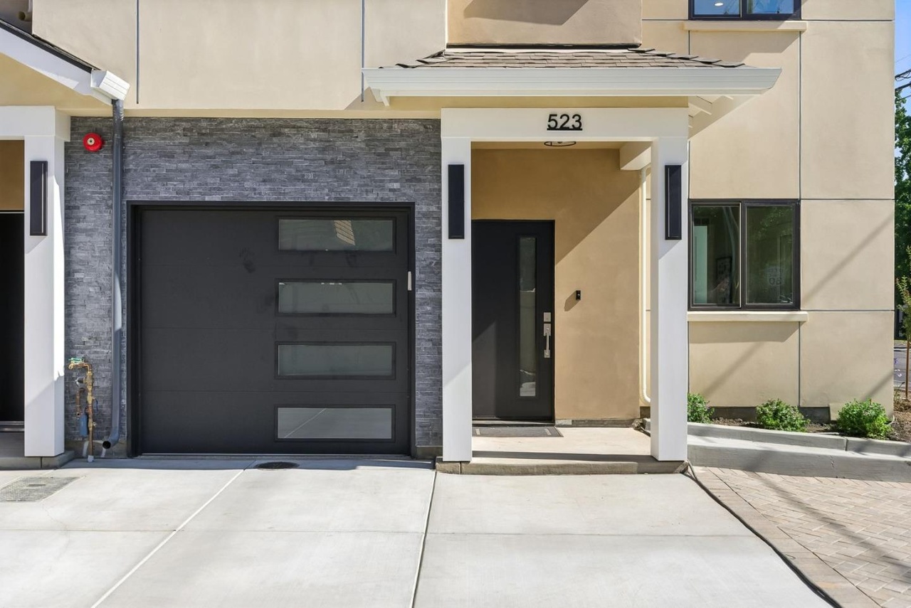 Covered front entry beside a dark garage door, with beige walls, white trim and gray exterior cladding.