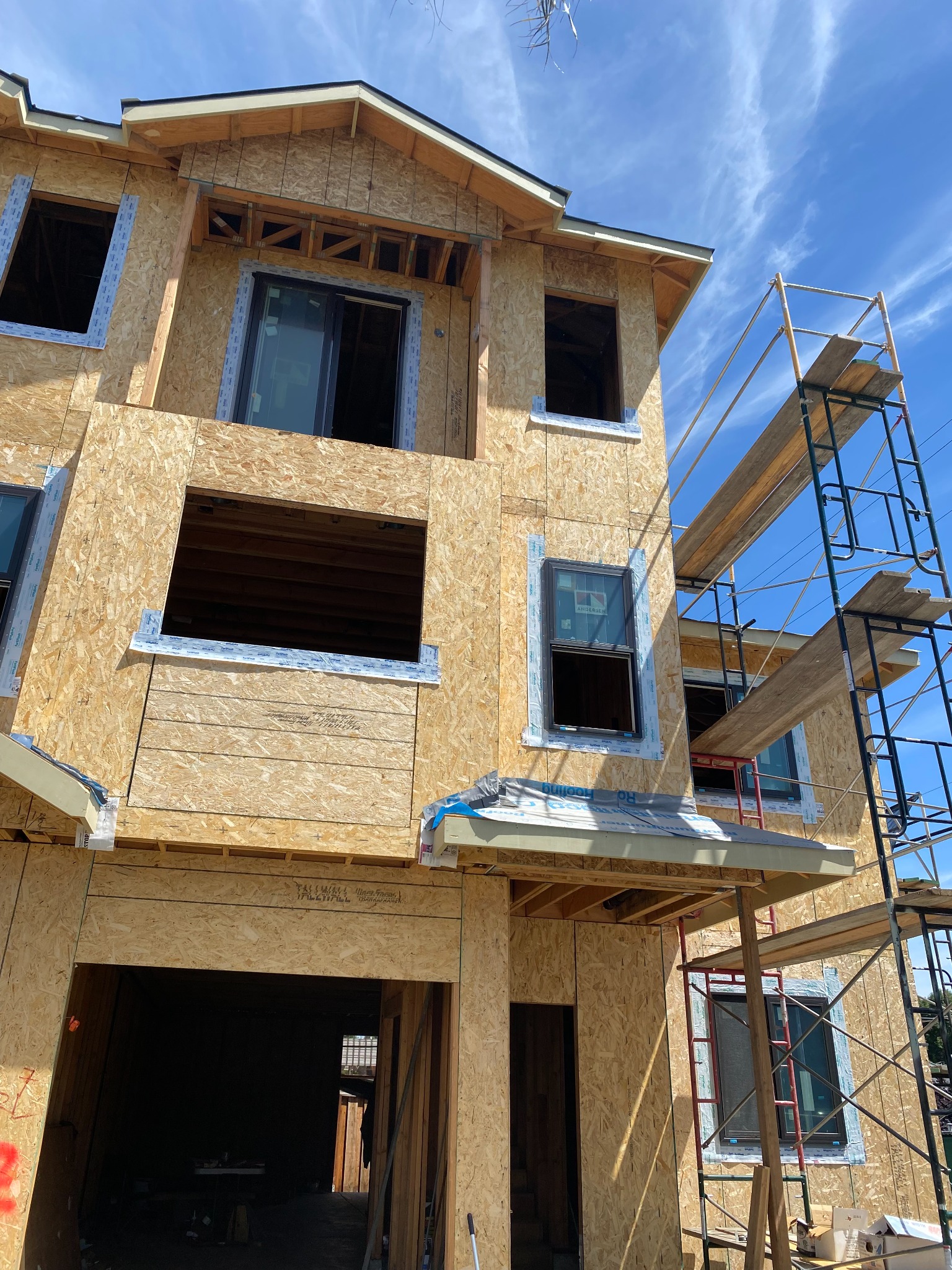 Multilevel building under construction with wood sheathing, window openings, roof overhangs and scaffolding.