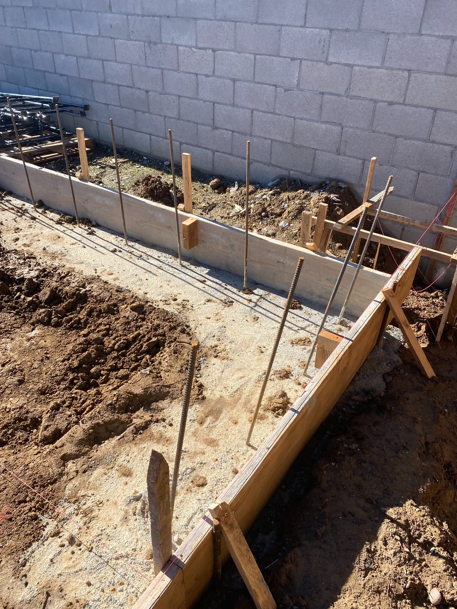 Excavated footing with timber forms and steel reinforcement beside a block wall.