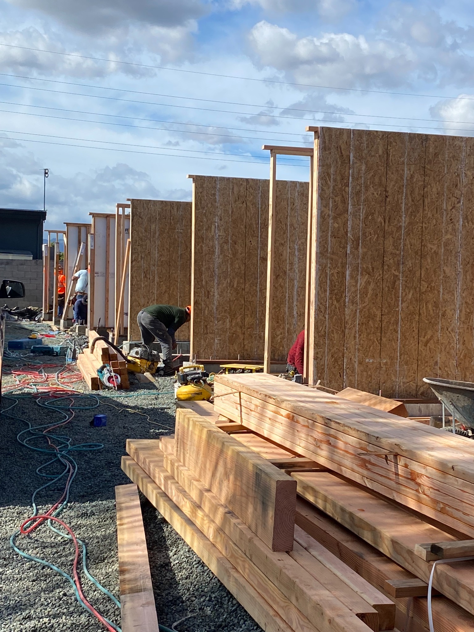 Wall framing and wood sheathing in progress, with stacked lumber and workers alongside the structure.